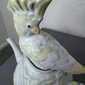 Może przedstawiać: Biały ceramiczny imbryk z pokrywką w kształcie papugi. Papuga ma ż&oacute;łte i niebieskie akcenty na grzebieniu i skrzydłach. Imbryk ma zakrzywioną wylewkę i uchwyt z kwiatowymi wzorami.