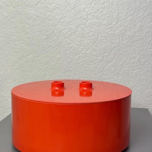 Op de afbeelding: Een feloranje, cilindervormige container met een plat, rond deksel. Twee kleine, cilindrische uitsteeksels bevinden zich op het deksel. De container is gemaakt van een glanzend materiaal en staat op een grijs oppervlak.