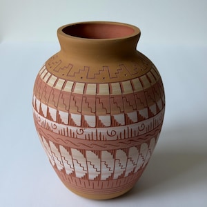 May include: A terracotta-colored ceramic vase with a rounded body and a narrow neck. The vase features a repeating geometric pattern in shades of cream and reddish-brown. The design includes stepped and wavy lines, creating a textured appearance.