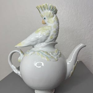 Może przedstawiać: Biały ceramiczny imbryk z okrągłym korpusem i zakrzywionym dziobkiem. Ozdobna, jasnoż&oacute;łta i biała figurka kakadu siedzi na pokrywce. Ptak ma grzebień i siedzi na kwiatowym wzorze. Imbryk ma uchwyt i stoi na małej podstawie.