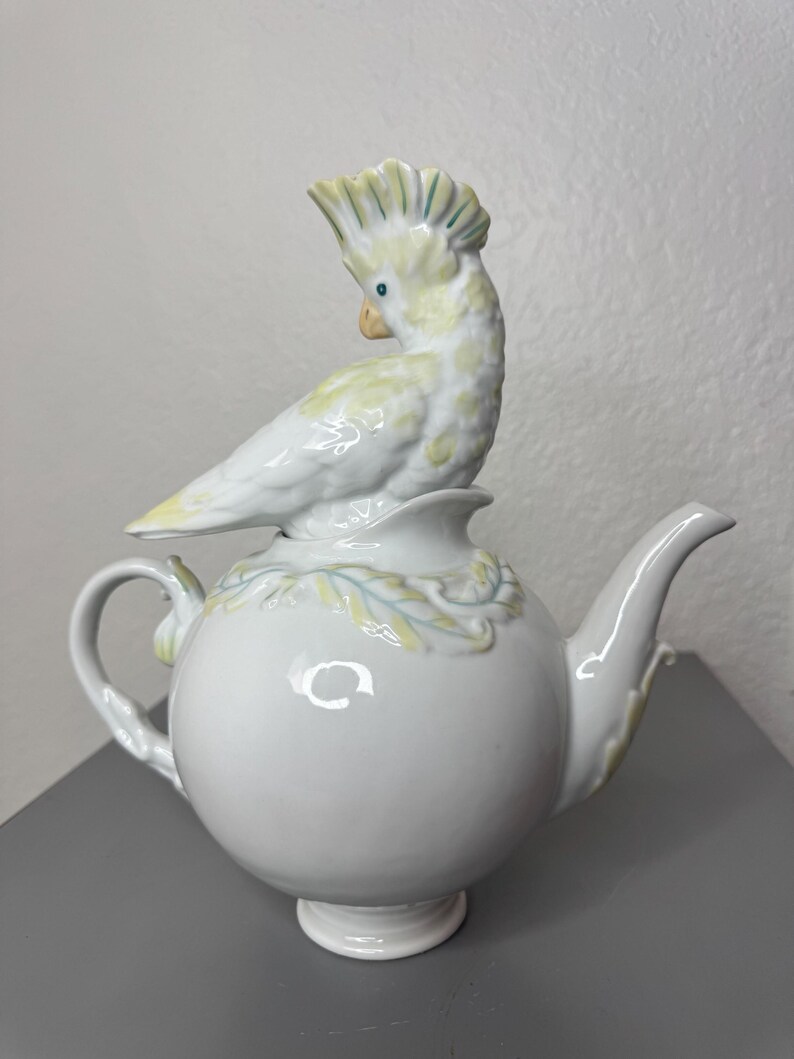 Może przedstawiać: Biały ceramiczny imbryk z okrągłym korpusem i zakrzywionym dziobkiem. Ozdobna, jasnoż&oacute;łta i biała figurka kakadu siedzi na pokrywce. Ptak ma grzebień i siedzi na kwiatowym wzorze. Imbryk ma uchwyt i stoi na małej podstawie.