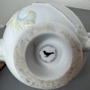 Może przedstawiać: Biały ceramiczny wazon z szeroką podstawą i zaokrąglonym korpusem. Wazon posiada subtelne ż&oacute;łte i niebieskie kwiatowe akcenty. Sp&oacute;d wazonu ma okrągłą pieczęć z sylwetką ptaka i napisem "Dishwasher Safe Made in China Fabriqu&eacute; en Chine."