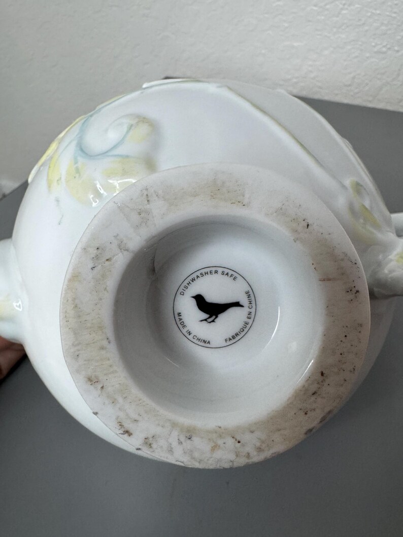 Może przedstawiać: Biały ceramiczny wazon z szeroką podstawą i zaokrąglonym korpusem. Wazon posiada subtelne ż&oacute;łte i niebieskie kwiatowe akcenty. Sp&oacute;d wazonu ma okrągłą pieczęć z sylwetką ptaka i napisem "Dishwasher Safe Made in China Fabriqu&eacute; en Chine."