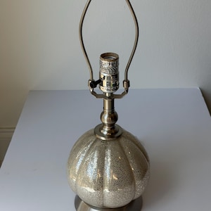 Peut inclure: Une lampe de table argentée avec une base ronde et texturée et un corps sphérique en forme de citrouille. La lampe a une armature métallique et une douille. La lampe est conçue pour une utilisation en intérieur et a un style classique et élégant.