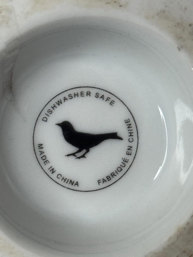 Może przedstawiać: Biały ceramiczny talerz z czarną sylwetką ptaka wewnątrz okrągłej obw&oacute;dki. Obw&oacute;dka zawiera tekst "Dishwasher Safe", "Made in China" i "Fabriqu&eacute; en Chine".