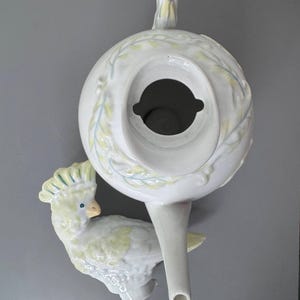 Może przedstawiać: Biały ceramiczny imbryk z motywem kakadu. Imbryk ma okrągły korpus, uchwyt i dzi&oacute;bek. Kakadu jest jasnoż&oacute;łty i biały z niebieskimi akcentami. Imbryk jest ozdobiony wzorami liści.
