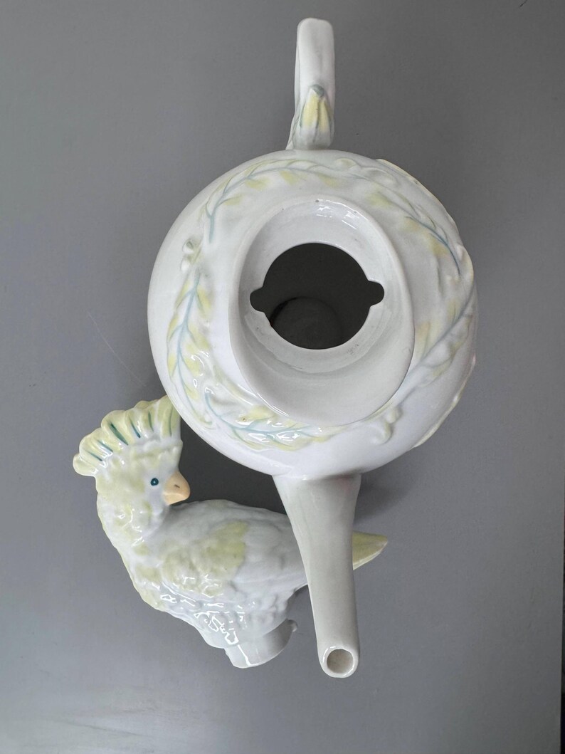 Może przedstawiać: Biały ceramiczny imbryk z motywem kakadu. Imbryk ma okrągły korpus, uchwyt i dzi&oacute;bek. Kakadu jest jasnoż&oacute;łty i biały z niebieskimi akcentami. Imbryk jest ozdobiony wzorami liści.