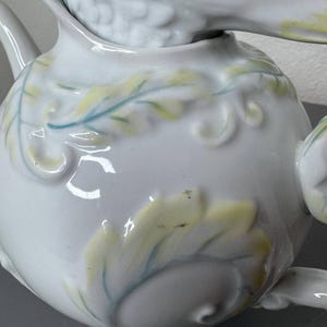 Może przedstawiać: Biały ceramiczny imbryk z pokrywką w kształcie ptaka. Imbryk posiada wytłaczane wzory liści i wir&oacute;w, zaakcentowane jasnoż&oacute;łtą i niebiesko-zieloną farbą. Widoczny jest dzi&oacute;bek i uchwyt, co sugeruje kompletny zestaw do herbaty.