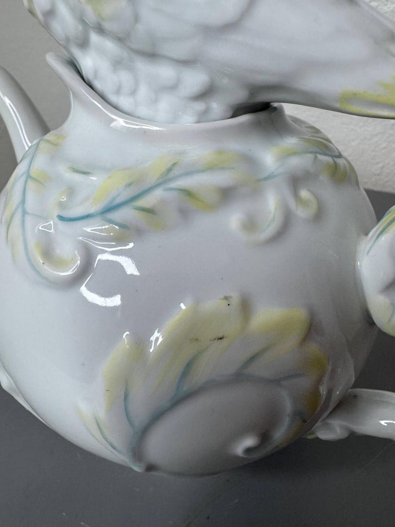 Może przedstawiać: Biały ceramiczny imbryk z pokrywką w kształcie ptaka. Imbryk posiada wytłaczane wzory liści i wir&oacute;w, zaakcentowane jasnoż&oacute;łtą i niebiesko-zieloną farbą. Widoczny jest dzi&oacute;bek i uchwyt, co sugeruje kompletny zestaw do herbaty.