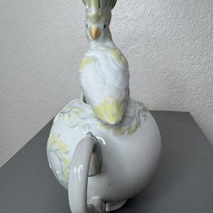 Może przedstawiać: Biały ceramiczny imbryk z pokrywką w kształcie papugi. Papuga jest biała z ż&oacute;łtymi i zielonymi akcentami. Imbryk ma okrągły korpus z zakrzywionym uchwytem i jest ozdobiony ż&oacute;łto-zielonymi wzorami kwiatowymi.