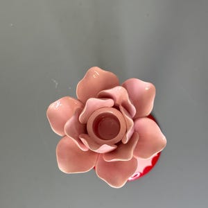 May include: A pink ceramic flower-shaped candle holder. The candle holder has a central well for a candle and is surrounded by multiple petals. The base is red with a white pattern. The overall design is decorative.