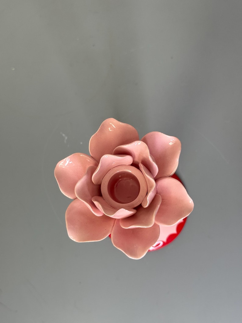 May include: A pink ceramic flower-shaped candle holder. The candle holder has a central well for a candle and is surrounded by multiple petals. The base is red with a white pattern. The overall design is decorative.