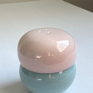 Könnte beinhalten: Ein zweiteiliges Salz- und Pfefferstreuer-Set aus Keramik. Das Oberteil hat eine hellrosa, kugelförmige Form mit drei Löchern. Das Unterteil hat eine hellblaue, kugelförmige Form. Das Set ist glänzend und steht auf einer weißen Oberfläche.