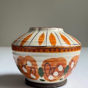 May include: A ceramic bowl with a lid. The cream-coloured base is decorated with orange floral patterns and green leaves. The lid has orange leaf-like designs and purple lines. The rim is decorated with a brown, wavy line.