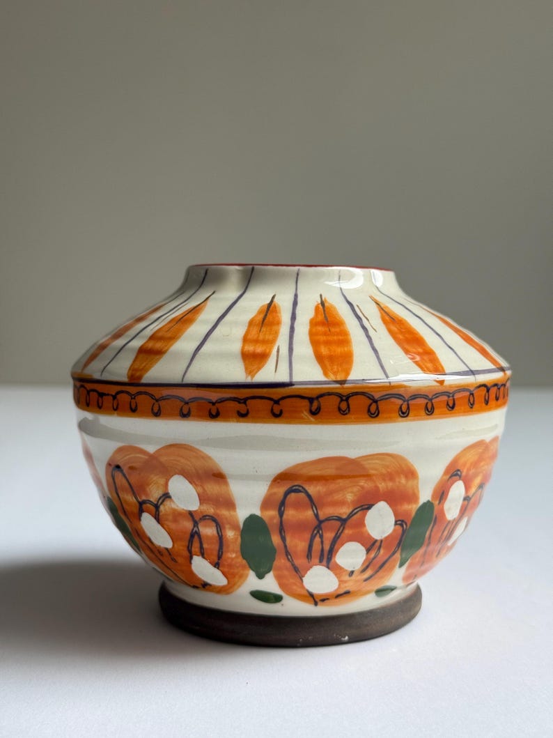 May include: A ceramic bowl with a lid. The cream-coloured base is decorated with orange floral patterns and green leaves. The lid has orange leaf-like designs and purple lines. The rim is decorated with a brown, wavy line.