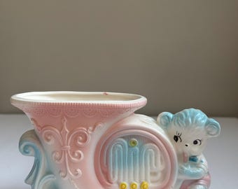 May include: A vintage ceramic planter in the shape of a French horn, painted in pastel pink, blue, and white. A small bear figure is attached to the side. The planter has a decorative design and three yellow accents.