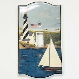 Peut inclure: Une plaque d'interrupteur rectangulaire ornée d'une scène côtière peinte représentant un phare rayé noir et blanc, un drapeau américain, un voilier sur une mer bleue et une colline herbeuse avec de petits bâtiments sous un ciel pâle.