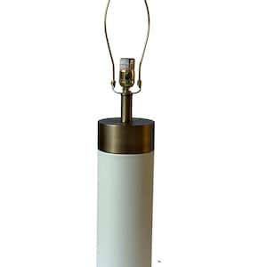 May include: A cylindrical table lamp with a white textured base and a brass-colored top and accents. The lamp features a gold-toned harp and bulb socket, ready for a light bulb. The design is modern and minimalist.