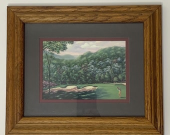 Puede incluir: Una impresión enmarcada de un paisaje de campo de golf. La obra representa a un golfista en un green, con trampas de arena y árboles en el fondo. El marco es de madera de tono medio, y la impresión está enmarcada con un passe-partout gris oscuro.