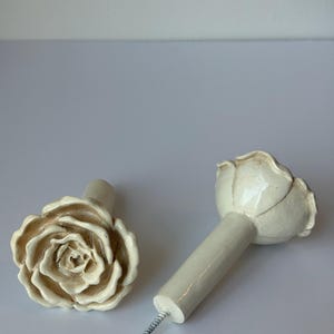 May include: Two off-white ceramic rose-shaped cabinet knobs. One knob is lying on its side, revealing the screw. The other knob is upright. The knobs have a detailed floral design and are suitable for furniture.