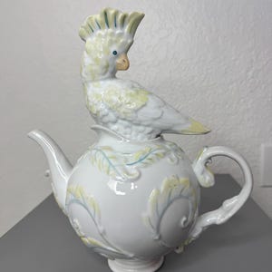Może przedstawiać: Biały ceramiczny imbryk z dekoracyjną papugą na g&oacute;rze. Imbryk posiada zakrzywioną wylewkę, szczeg&oacute;łowy uchwyt i jest ozdobiony ż&oacute;łtymi i niebieskimi wzorami liści. Papuga ma ż&oacute;łte i niebieskie akcenty.