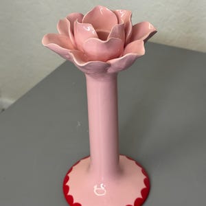 May include: A pink ceramic candle holder in the shape of a rose. The holder features a long stem and a wide base with a red, scalloped edge. The rose petals are intricately detailed and layered, creating a realistic look.