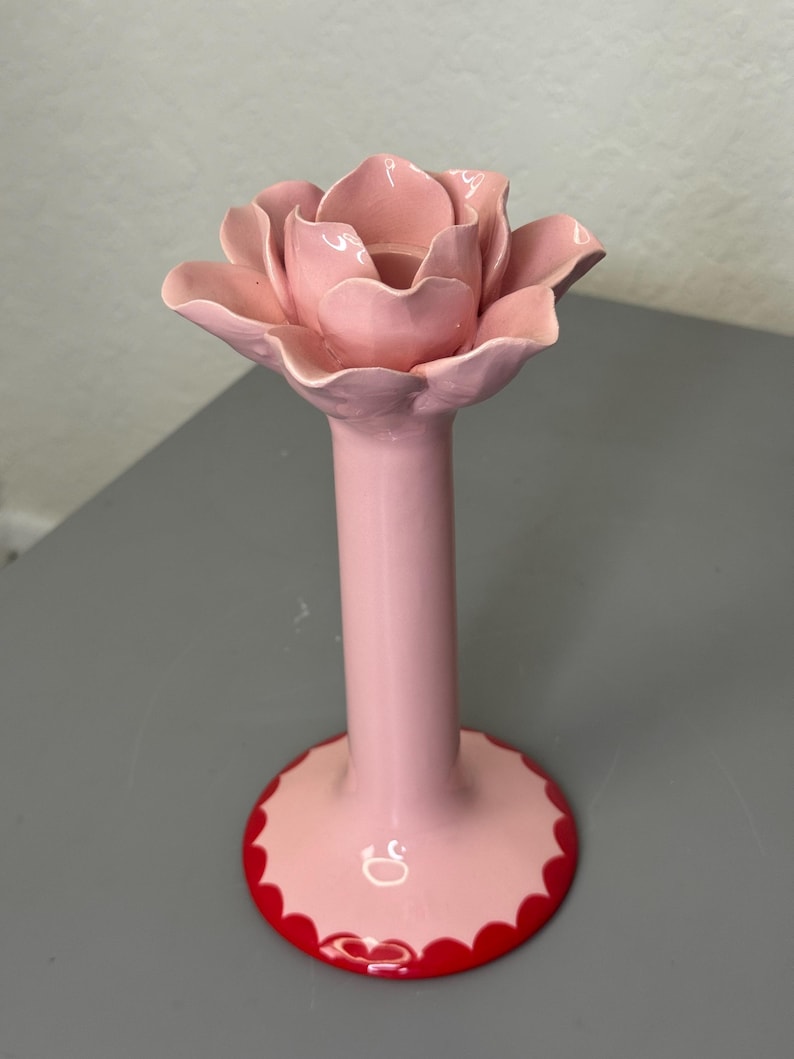 May include: A pink ceramic candle holder in the shape of a rose. The holder features a long stem and a wide base with a red, scalloped edge. The rose petals are intricately detailed and layered, creating a realistic look.