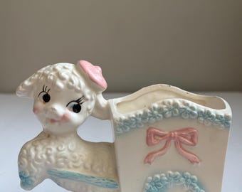 May include: A vintage ceramic lamb planter in white with a pink bow and blue accents. The lamb is pulling a cart decorated with a pink bow and blue floral details. The planter is designed for small plants or flowers.