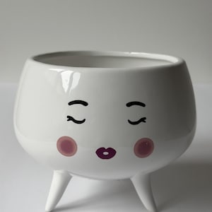 May include: A white ceramic planter with a whimsical face design. The planter features closed eyes, rosy cheeks, and a purple lip design. It stands on three small, white legs, adding a touch of playful charm to any space.