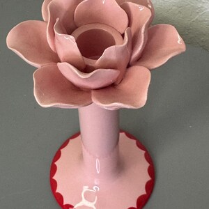 May include: A pink ceramic rose-shaped candle holder. The rose has multiple petals and a central well for a candle. The base is pink with a red scalloped edge. The candle holder is on a grey surface.