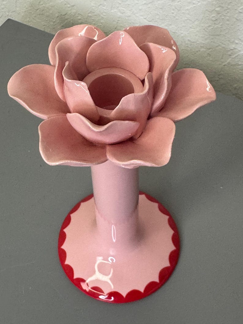 May include: A pink ceramic rose-shaped candle holder. The rose has multiple petals and a central well for a candle. The base is pink with a red scalloped edge. The candle holder is on a grey surface.