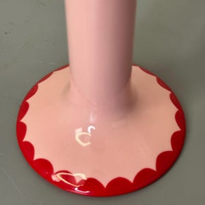 May include: A close-up of a pink, decorative stand with a scalloped red border. The stand has a long, cylindrical stem and a circular base. The base is light pink with a bright red, scalloped edge.