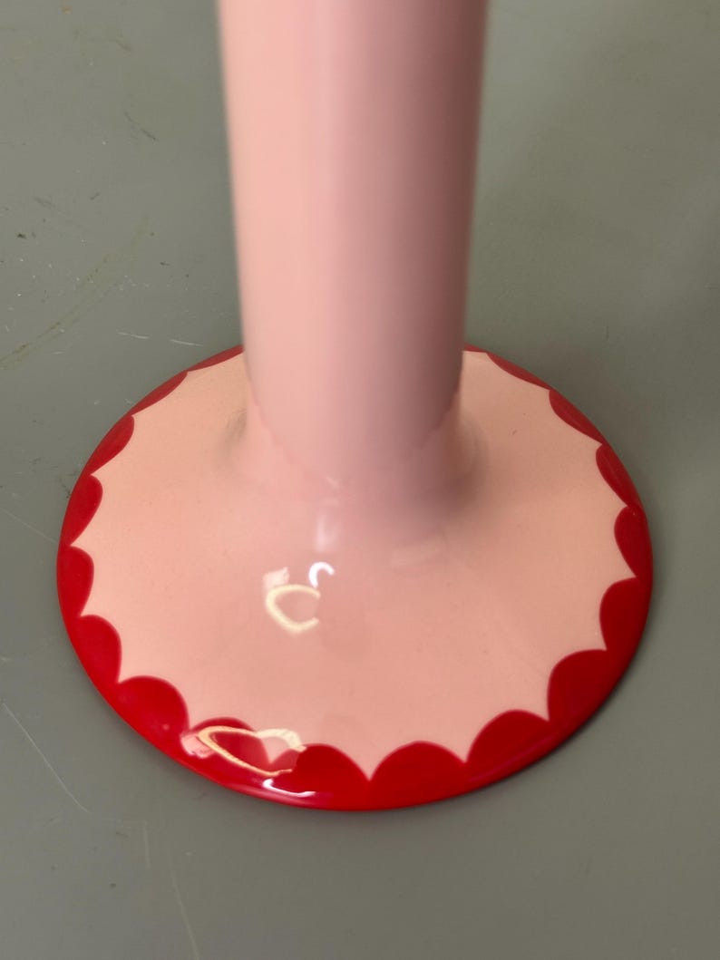 May include: A close-up of a pink, decorative stand with a scalloped red border. The stand has a long, cylindrical stem and a circular base. The base is light pink with a bright red, scalloped edge.
