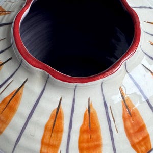 May include: A ceramic vase with a wide, fluted rim and a dark interior. The exterior is off-white with orange leaf-like designs and vertical purple stripes. The rim is outlined in red.