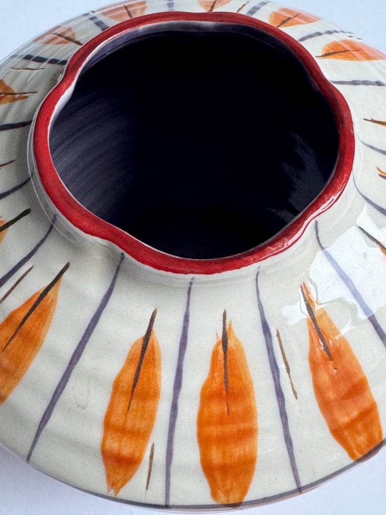 May include: A ceramic vase with a wide, fluted rim and a dark interior. The exterior is off-white with orange leaf-like designs and vertical purple stripes. The rim is outlined in red.