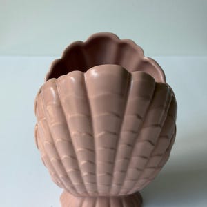 May include: A dusty rose ceramic planter shaped like a large scallop shell. The vessel features a scalloped rim and a textured exterior with vertical ridges radiating from the base to the top edge, creating a shell-like appearance.