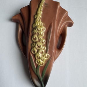 May include: A brown ceramic wall pocket vase with a stylized floral design. The vase features a central yellow flower stalk with green leaves, set against a textured brown background. The vase has a unique shape with two side handles.