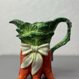 May include: A ceramic pitcher shaped like a carrot, with green leafy top and a white bow with a green plaid pattern. The pitcher has an orange base and a curved handle.
