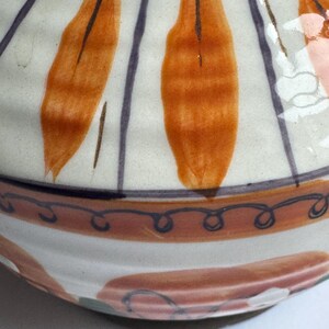 May include: A ceramic bowl with a cream exterior, decorated with orange leaf-like shapes and dark purple vertical stripes. The rim has a brown band with a blue swirl design. The base features a pink and white pattern.