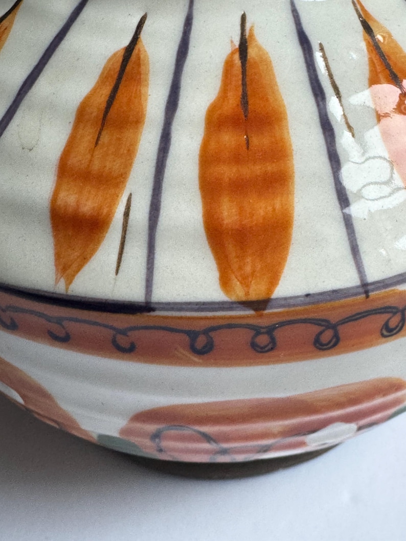 May include: A ceramic bowl with a cream exterior, decorated with orange leaf-like shapes and dark purple vertical stripes. The rim has a brown band with a blue swirl design. The base features a pink and white pattern.