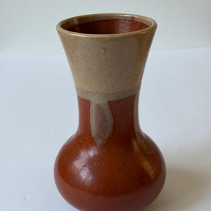 May include: A ceramic vase with a bulbous base and a flared rim. The lower portion features a glossy, deep reddish-brown glaze, while the upper section is finished in a speckled, light beige glaze that drips down onto the darker base color.