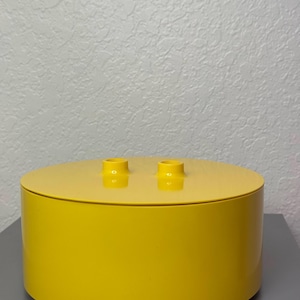May include: A bright yellow, cylindrical container with a flat, round lid. Two small, yellow cylindrical protrusions are on the lid. The container is sitting on a gray surface against a white wall.