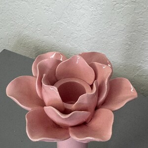 May include: A pink ceramic candle holder in the shape of a rose. The candle holder has a round base and a cylindrical stem supporting the flower-shaped top. The petals are detailed and the colour is a soft, muted pink.