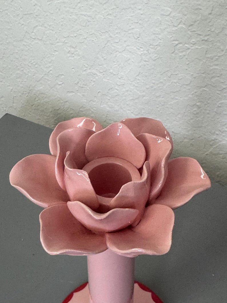May include: A pink ceramic candle holder in the shape of a rose. The candle holder has a round base and a cylindrical stem supporting the flower-shaped top. The petals are detailed and the colour is a soft, muted pink.