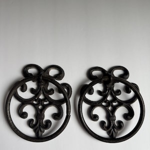Puede incluir: Dos tiradores de cajón ornamentados de color bronce oscuro. Cada tirador presenta un asa circular con un diseño intrincado y simétrico en el interior. La parte superior de cada tirador tiene un elemento decorativo en forma de lazo.