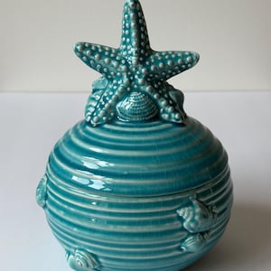 Può includere: Un barattolo in ceramica turchese con coperchio a forma di stella marina. Il barattolo ha un corpo rotondo e scanalato con accenti di conchiglie. Il coperchio a forma di stella marina è strutturato con piccoli rilievi e una conchiglia alla base. È un oggetto decorativo.