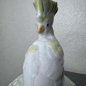 Może przedstawiać: Ceramiczna figurka ptaka kakadu. Ptak jest gł&oacute;wnie biały z ż&oacute;łtymi i zielonymi akcentami na czubku i skrzydłach. Ma ż&oacute;łty dzi&oacute;b i małe czarne oczy. Figurka jest szczeg&oacute;łowa i wydaje się być ręcznie malowana.