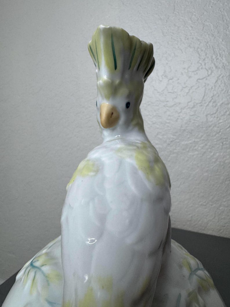 Może przedstawiać: Ceramiczna figurka ptaka kakadu. Ptak jest gł&oacute;wnie biały z ż&oacute;łtymi i zielonymi akcentami na czubku i skrzydłach. Ma ż&oacute;łty dzi&oacute;b i małe czarne oczy. Figurka jest szczeg&oacute;łowa i wydaje się być ręcznie malowana.