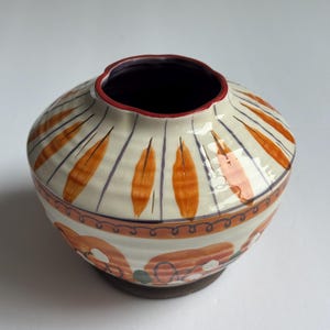 May include: A ceramic vase with a wide, rounded body and a narrow opening. The vase is cream-coloured with orange, purple, and red painted designs. The base is a dark brown colour. The vase is likely handmade and suitable for home decor.