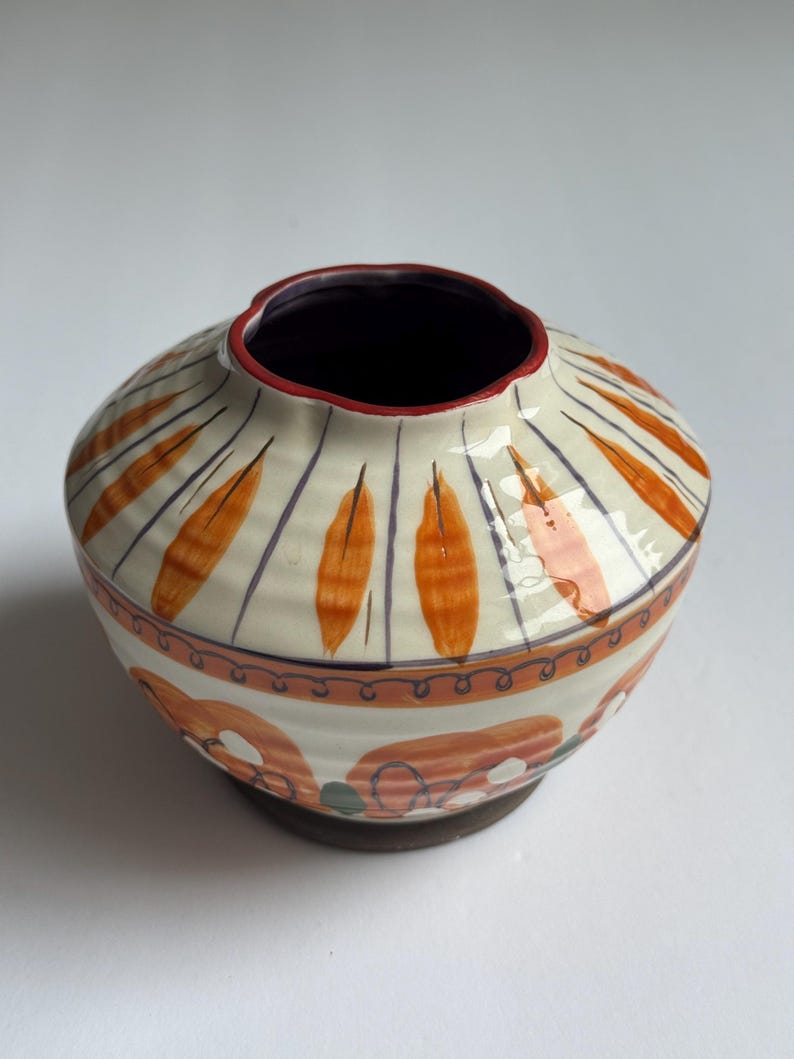 May include: A ceramic vase with a wide, rounded body and a narrow opening. The vase is cream-coloured with orange, purple, and red painted designs. The base is a dark brown colour. The vase is likely handmade and suitable for home decor.